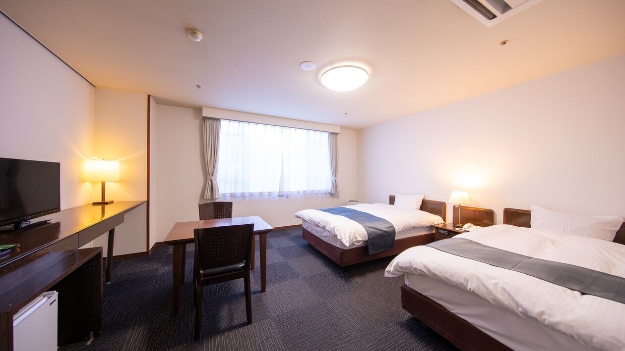 Ein Hotelzimmer mit zwei Betten, einem Schreibtisch, einem Sessel, einem Fernseher und einem Fenster mit Vorhängen.