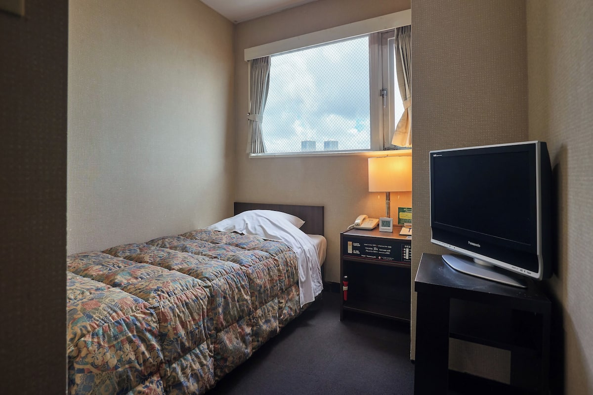 A hotel room with a single bed, a bedside table with a lamp, a television, and a window with curtains.