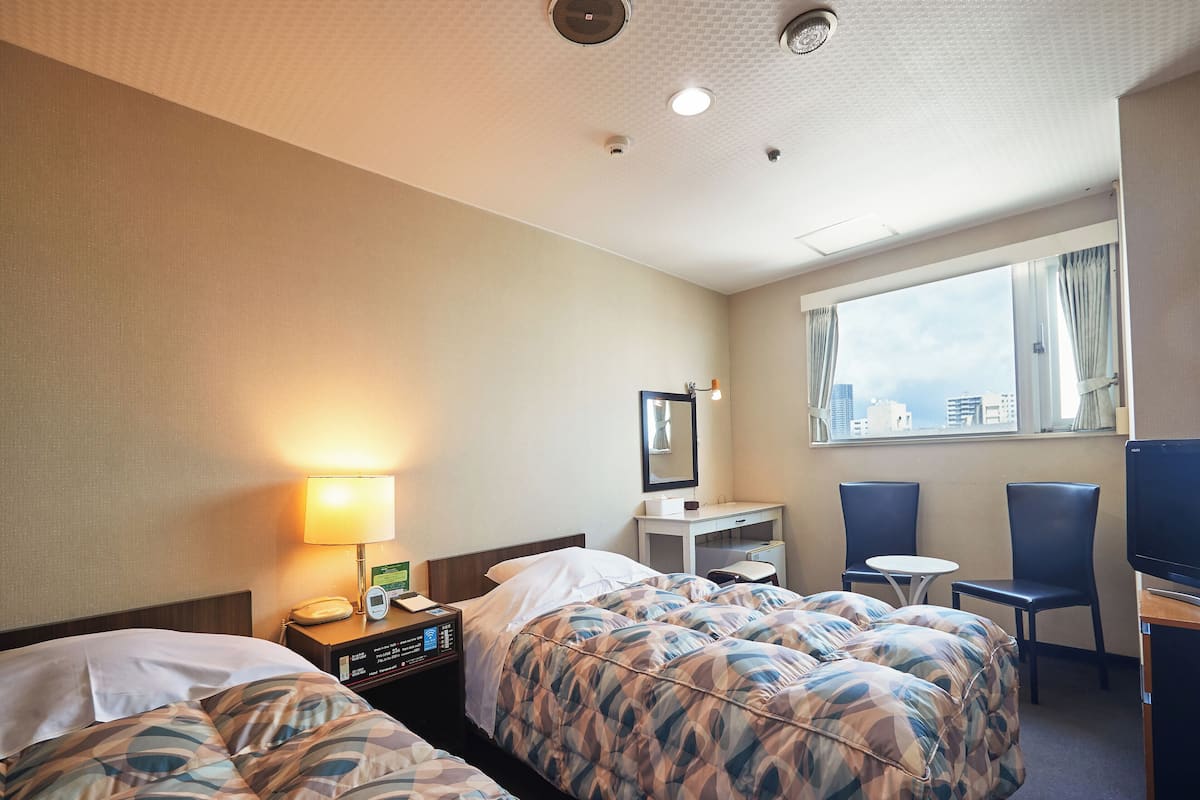A hotel room with two beds, a desk, a chair, a TV, and a window with a city view.