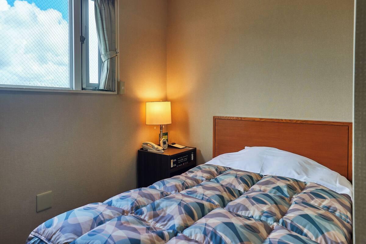 A hotel room with a bed, a bedside table with a lamp, a phone, and a window with curtains.