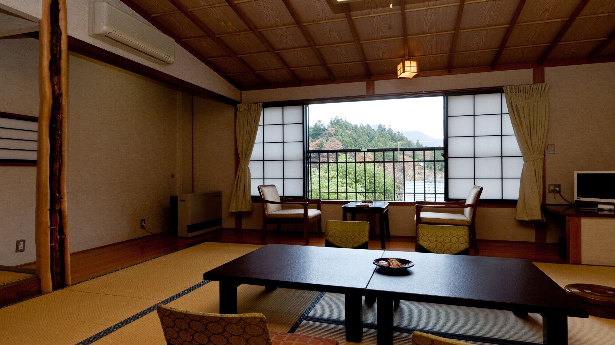 Una habitación japonesa tradicional con suelo de tatami, una mesa baja, sillas, un televisor y vistas a zonas verdes.