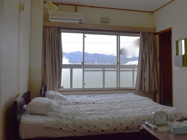 Habitación de hotel con cama, mesita de noche con reloj, ventana con cortinas, aire acondicionado y vista al agua.