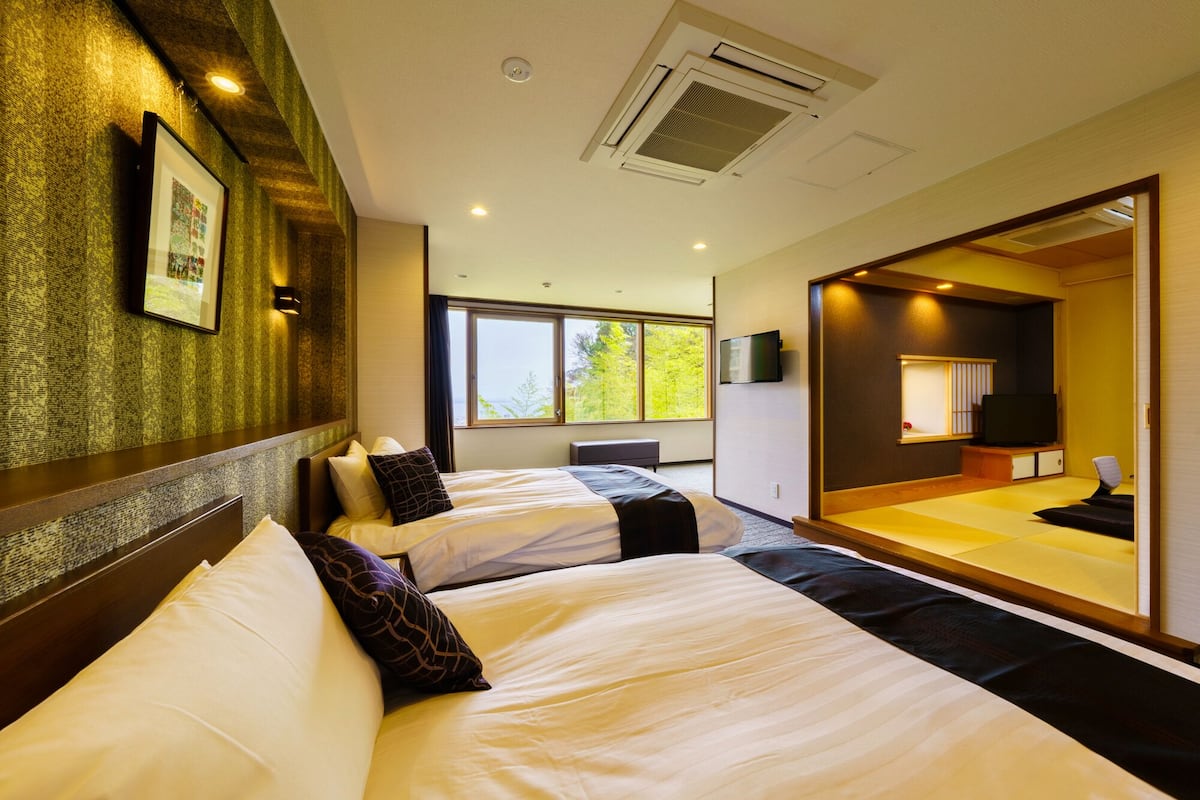 Habitación de hotel con dos camas, un espejo grande, televisión y vistas a árboles por la ventana.