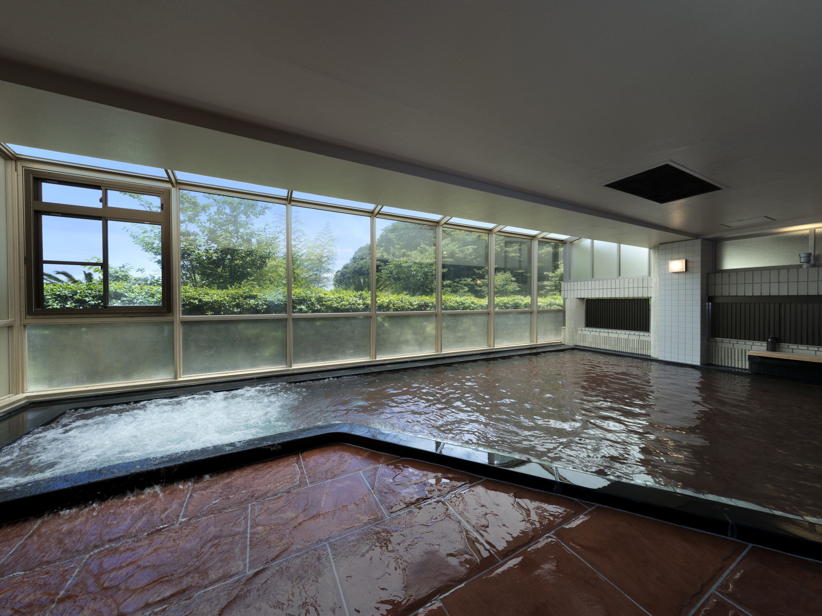 A spacious indoor area with a large window overlooking greenery, a wet floor indicating recent cleaning, and a tiled floor.