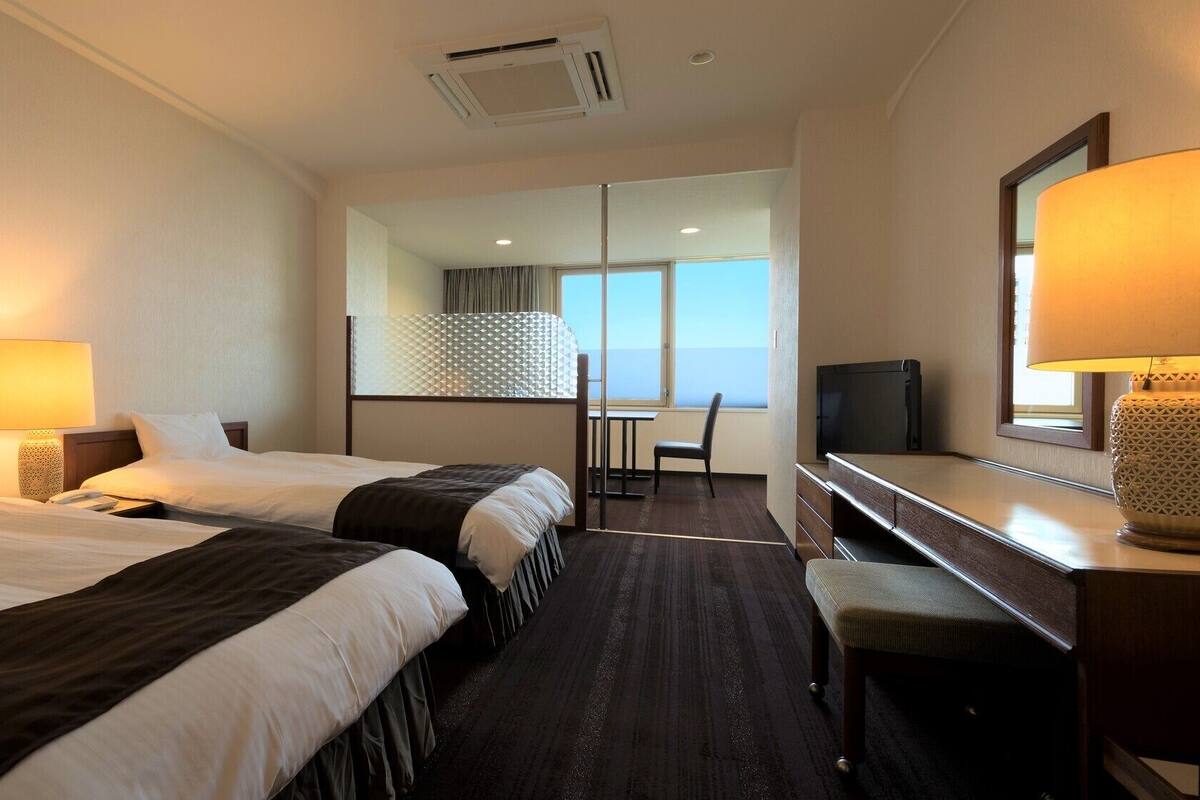 Habitación de hotel con dos camas, un escritorio, una silla, un televisor y un ventanal grande.
