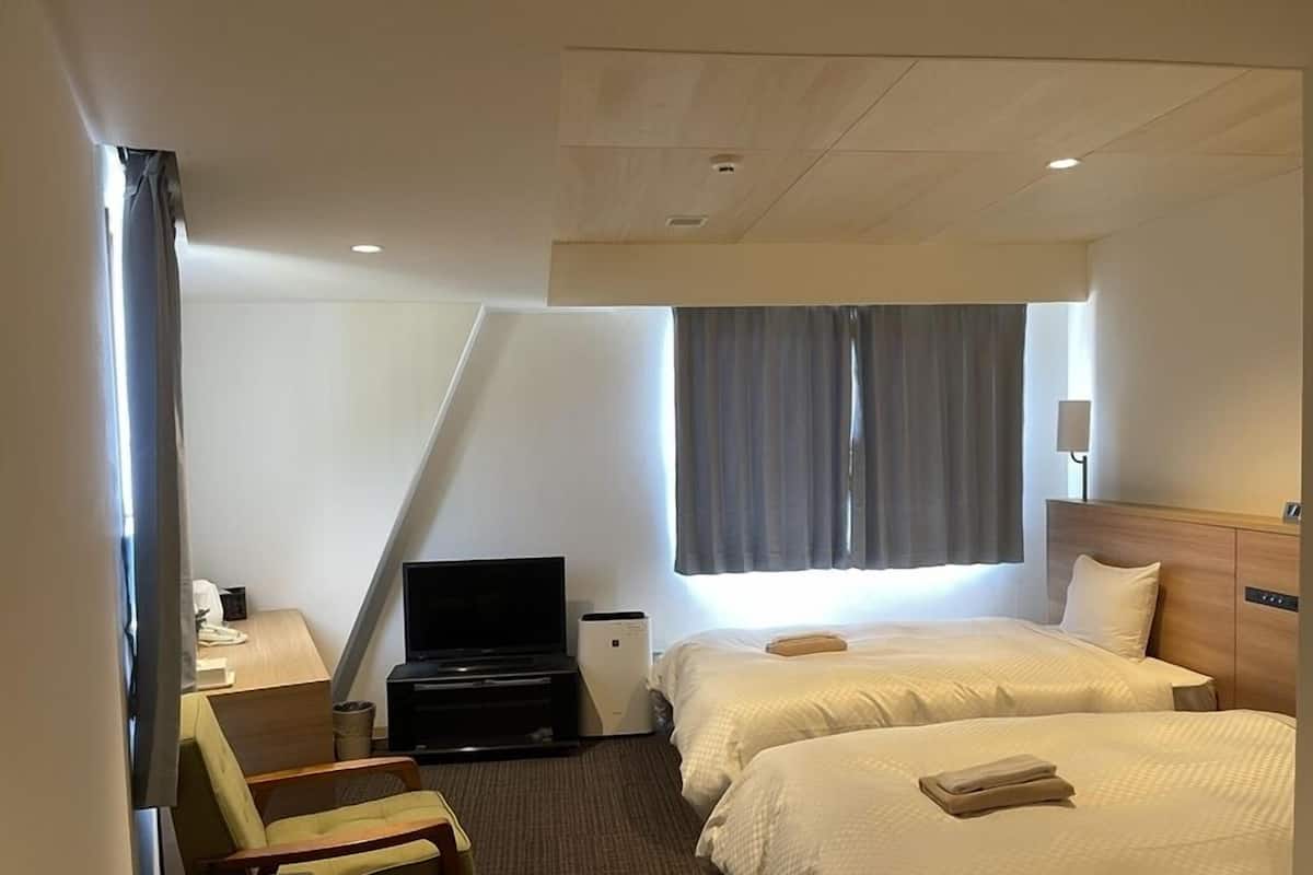 Ein Hotelzimmer mit zwei Betten, einem Schreibtisch mit Stuhl, einem Fernseher und einem Fenster mit Vorhängen.