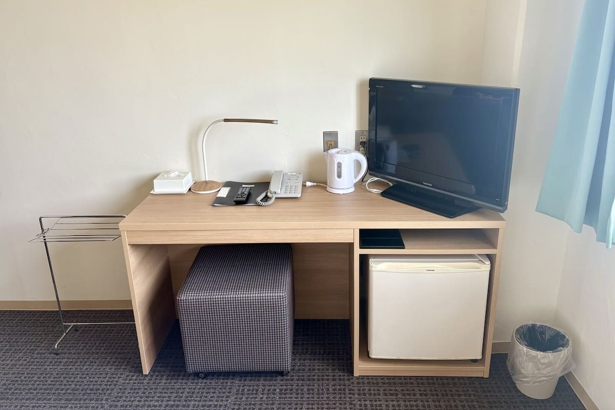 Ein Hotelzimmer mit einem Holztisch, einem Flachbildfernseher, einer Schreibtischlampe, einem Telefon, einer Kaffeemaschine, einem kleinen Kühlschrank und einem Mülleimer.