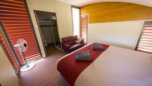 Quarto de hotel com cama, tapete vermelho, ventilador, sofá vermelho e banheiro visível através de uma porta aberta.