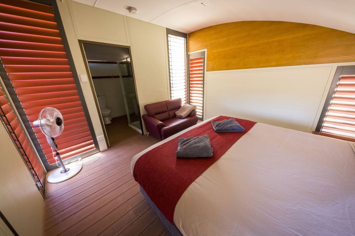 Habitación de hotel con cama, alfombra roja, ventilador, sofá rojo y baño visible a través de una puerta abierta.
