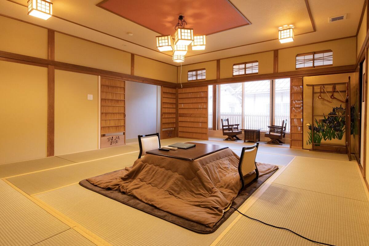 Une pièce avec un sol recouvert de tatamis, une petite table, des chaises et un luminaire de style japonais traditionnel au plafond.
