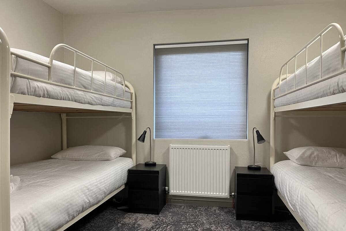 A room with two bunk beds, a radiator, a window with blinds, and two black speakers.