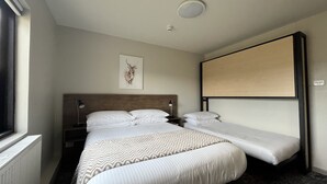 A hotel room with a large bed, a smaller bed, a headboard, a nightstand, a lamp, a ceiling light, a window, and a wall-mounted TV. - Swagman Chalet (Perisher Valley)