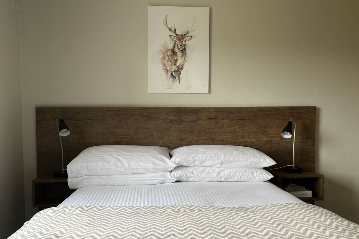 A neatly made bed with a patterned blanket, two bedside lamps, and a framed artwork of a deer on the wall.