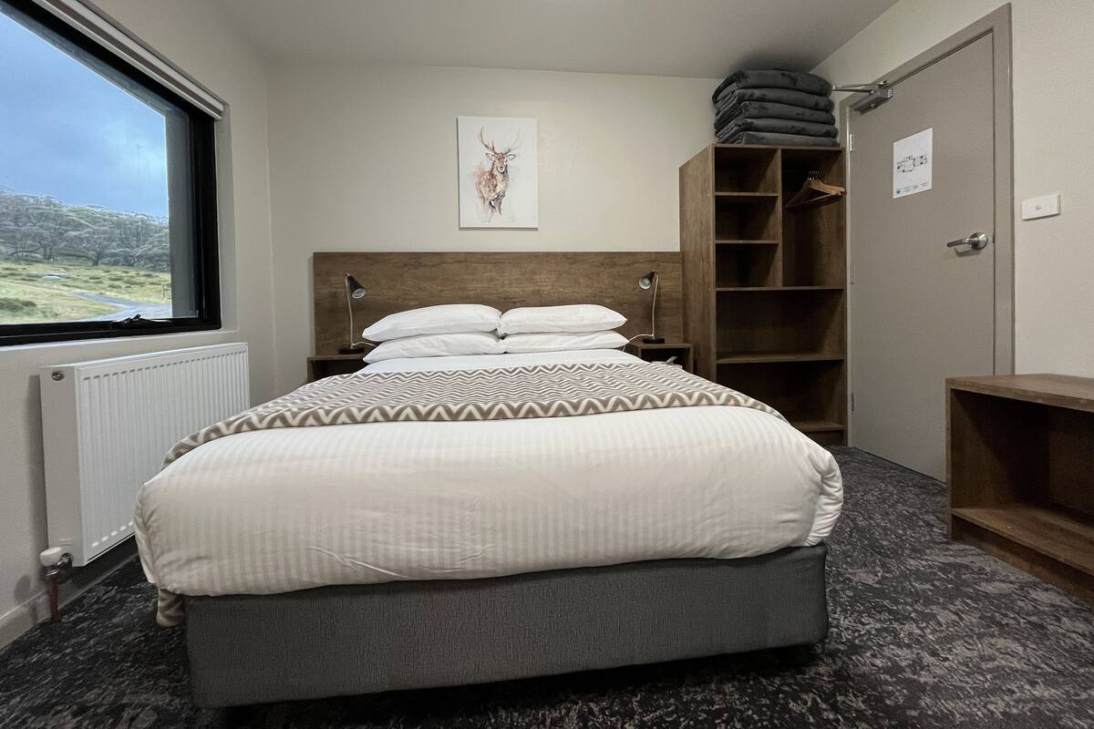 A bedroom with a bed, a window, a radiator, a headboard, a bedside table, a lamp, a wall-mounted artwork, a wardrobe, and a door with a sign.