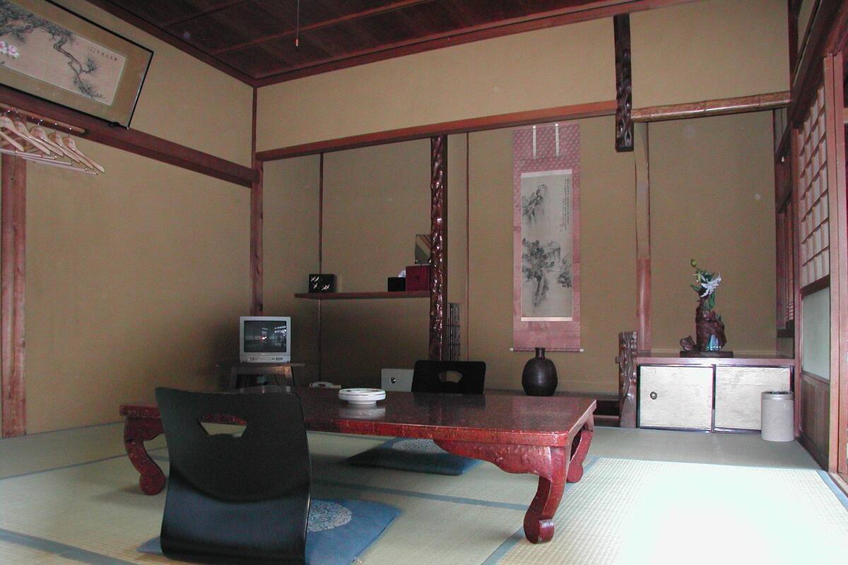 Une pièce de style japonais traditionnel, avec un sol recouvert de tatamis, une table basse, une chaise noire, un téléviseur et un vase décoratif.