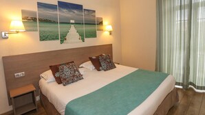 A neatly made bed with a green bedspread, two patterned pillows, and a wooden headboard. There are two wall-mounted lamps on either side of the bed, and a small nightstand to the left. - Hotel Duc de Padoue (Corte)
