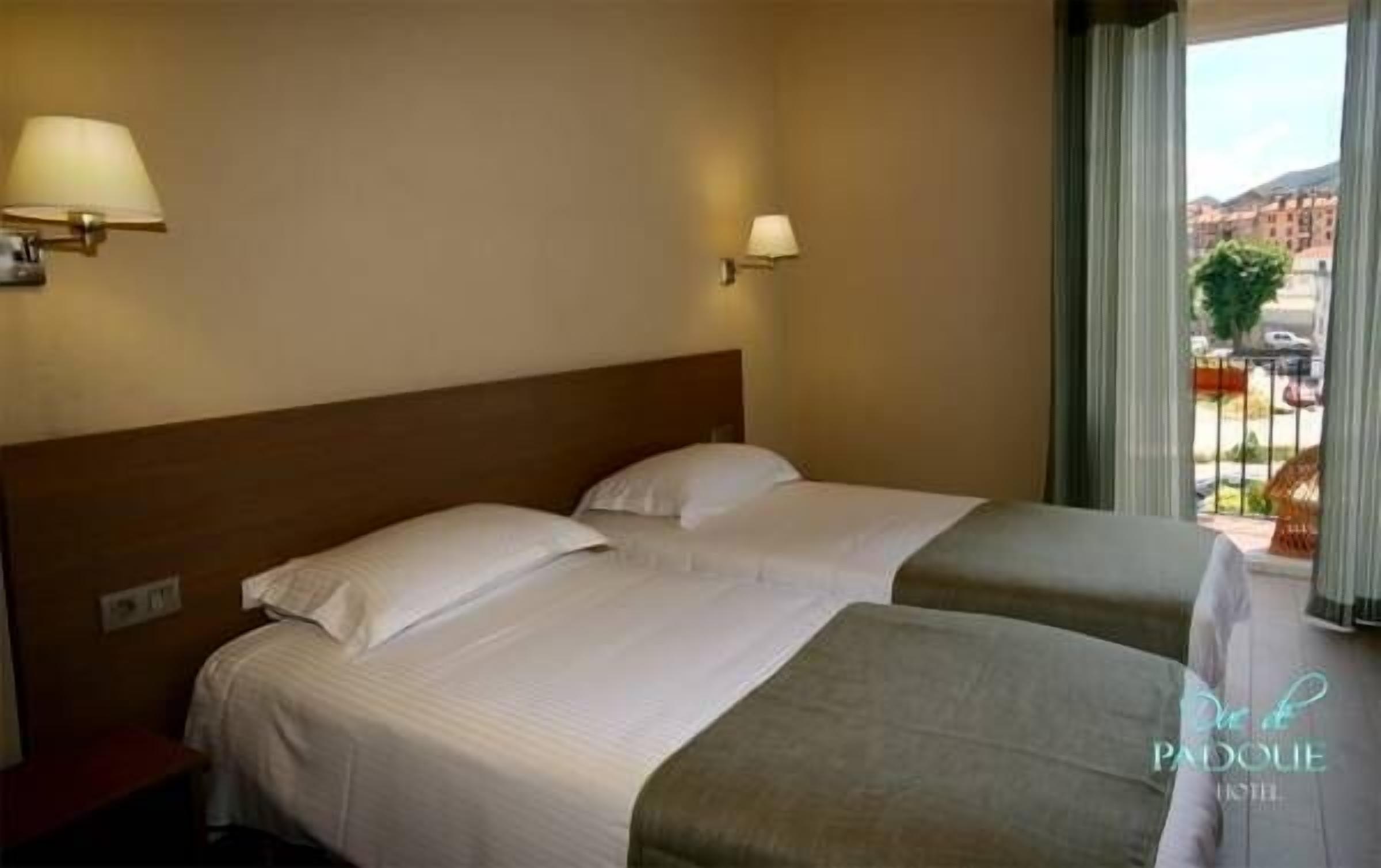A hotel room with two beds, a wooden headboard, a window with a view of buildings, and a lamp on the wall.