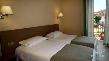 A hotel room with two beds, a wooden headboard, a window with a view of buildings, and a lamp on the wall.