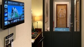 A hotel reception area with a digital screen displaying hotel information, a desk with a lamp, and a blue carpeted entrance. - Hotel Duc de Padoue (Corte)