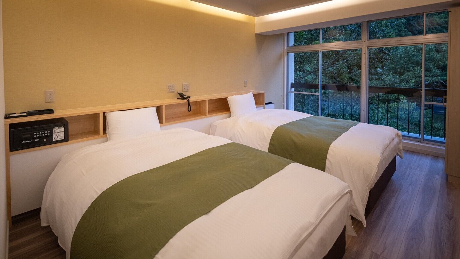 A hotel room with two beds, a bedside table with a phone, a radio, and a window with a view of trees.