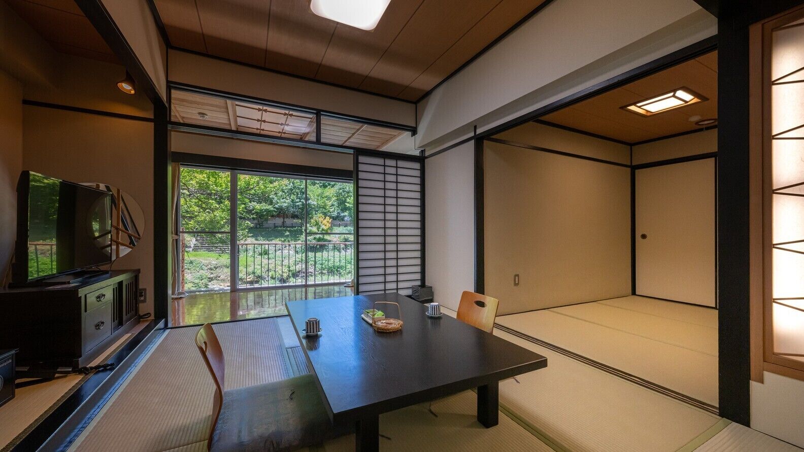 Una habitación japonesa tradicional con suelo de tatami, una mesa baja, un televisor y una puerta corrediza que da a un balcón con vistas a la vegetación.