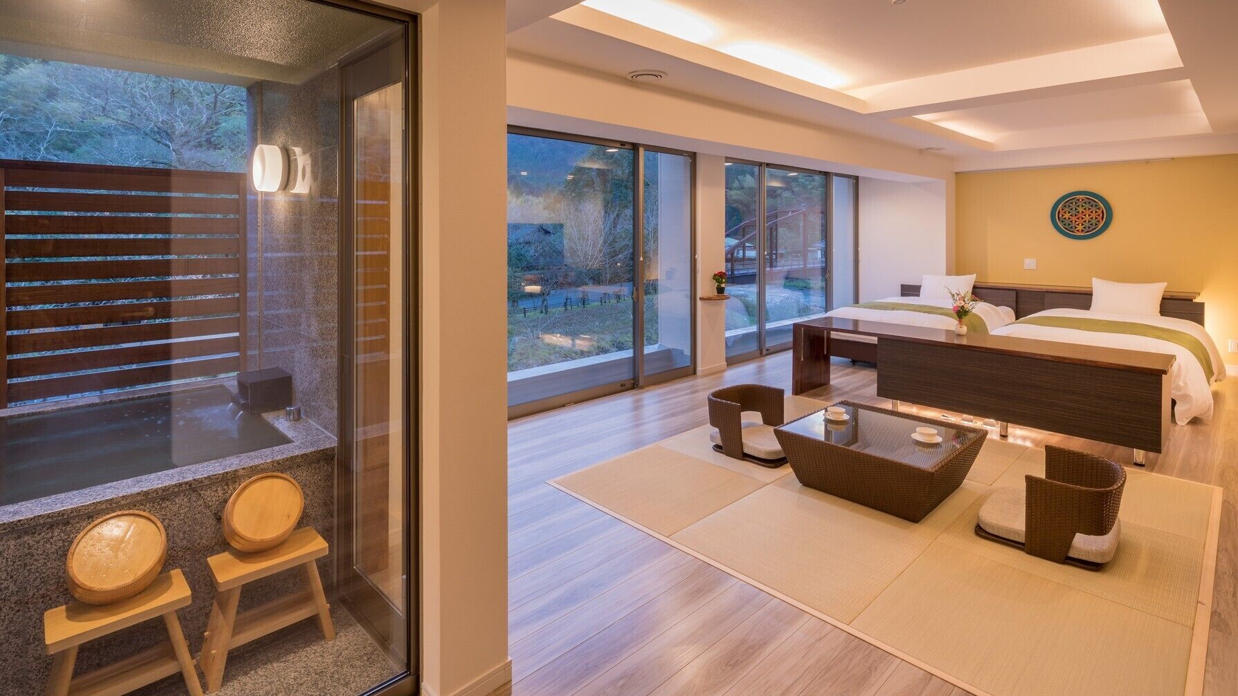 A modern hotel room with a large bed, a sitting area with a coffee table, and a view of the outdoors through large windows.