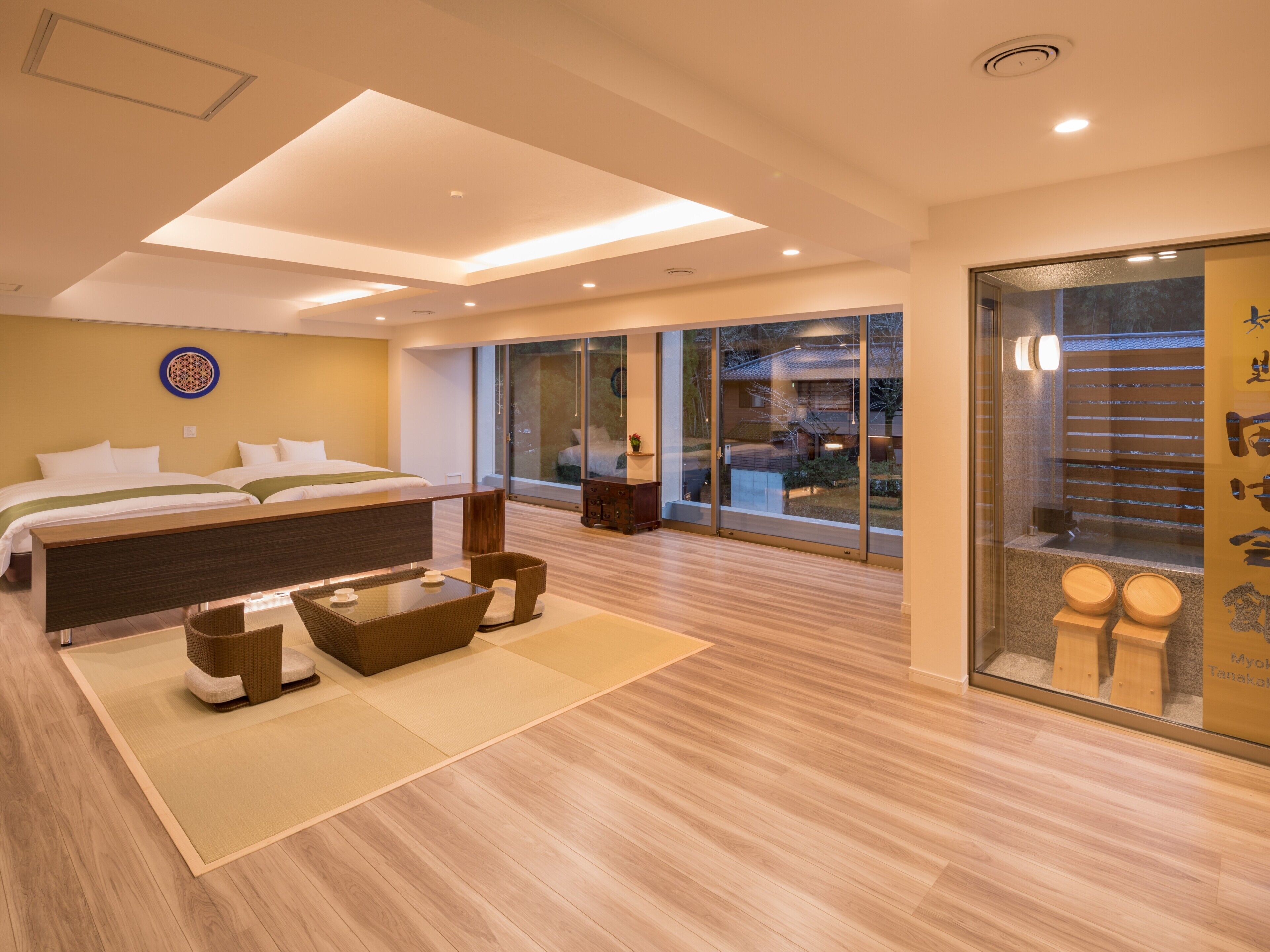 A modern hotel room with a large bed, a sitting area with a table and chairs, a large sliding glass door leading to a balcony, and a view of the outside.
