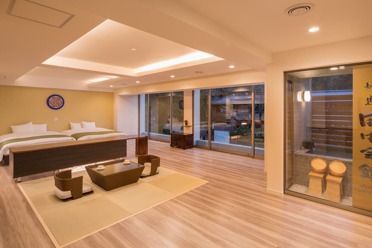 A modern hotel room with a large bed, a sitting area with a table and chairs, a large sliding glass door leading to a balcony, and a view of the outside.