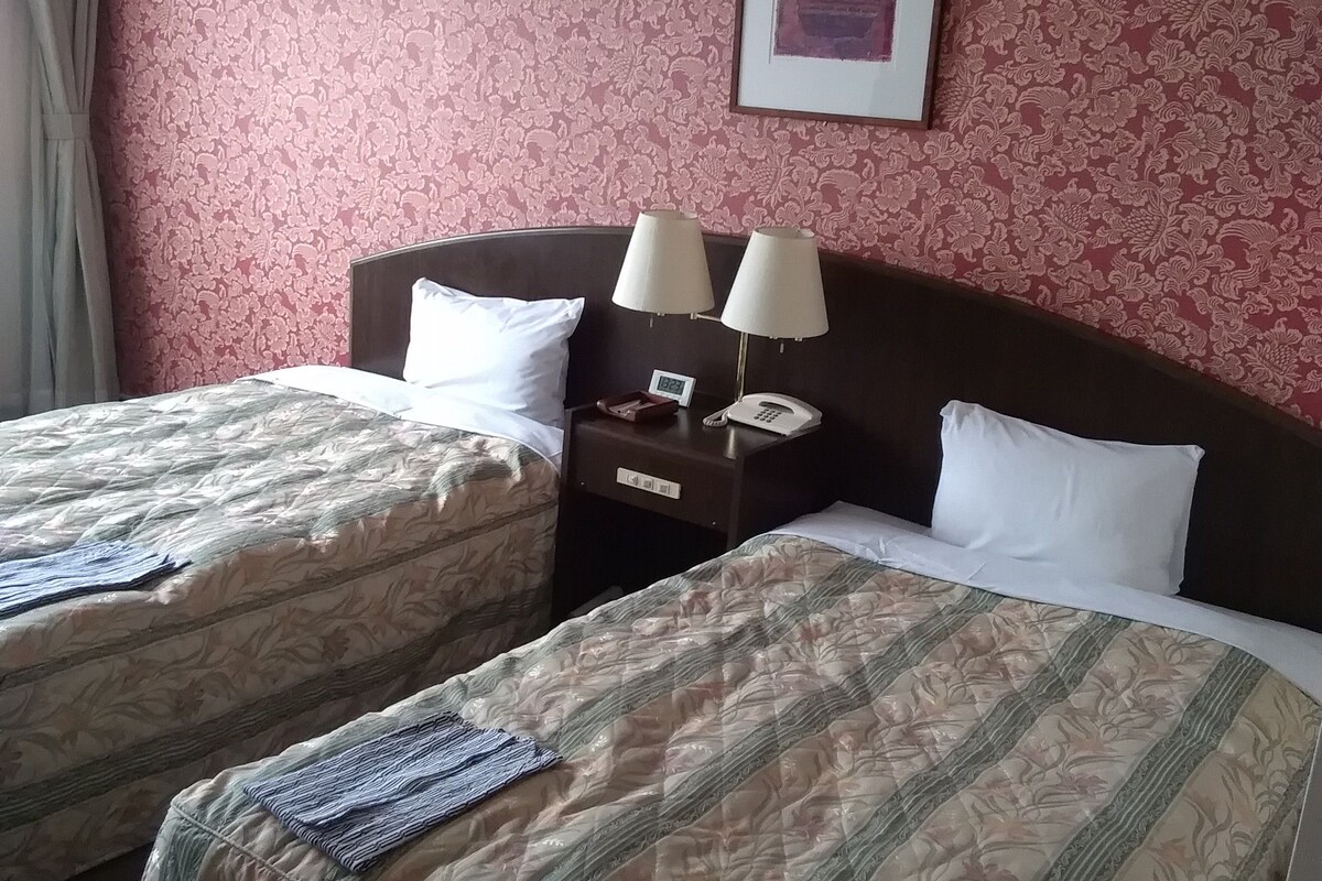 Een hotelkamer met twee bedden, een nachtkastje met lampen, een klok en een lijst aan de muur.