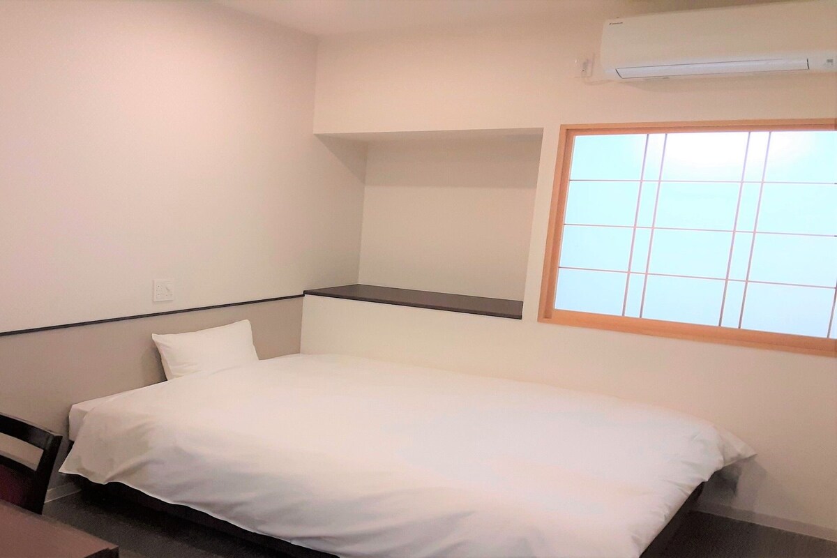 Een kleine eenpersoonskamer met een bureau en stoel, een raam met zonwering en een wandairconditioner.