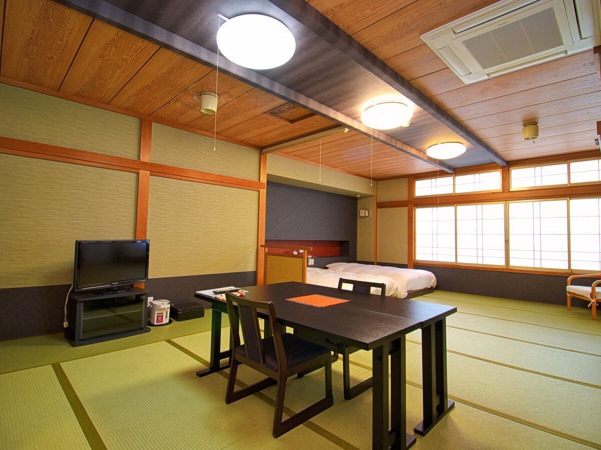 A traditional Japanese-style room with tatami flooring, a low table with chairs, a bed, a television, and a large window.