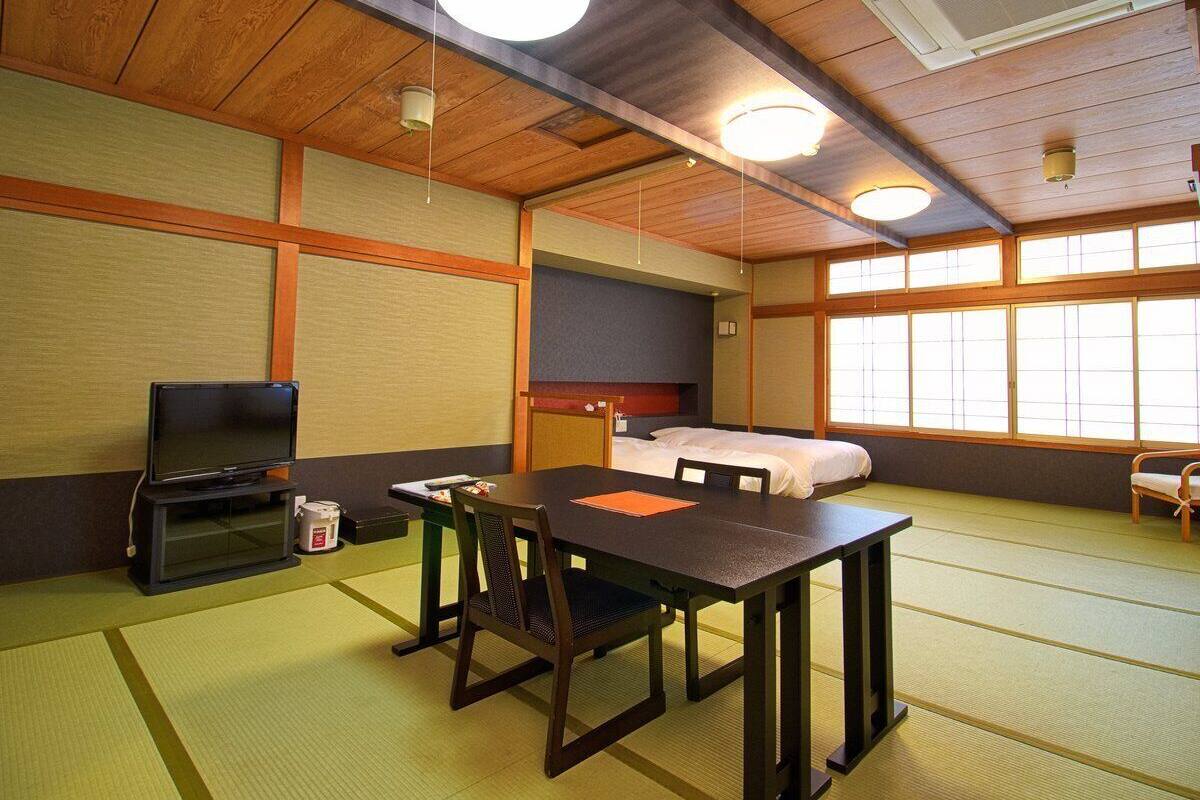 Ein traditioneller japanischer Raum mit Tatami-Böden, einem niedrigen Tisch mit Stühlen, einem Bett, einem Fernseher und einem großen Fenster.