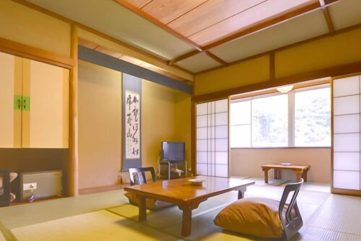 Ein traditioneller japanischer Raum mit Tatami-Bodenbelag, einem niedrigen Tisch, Stühlen, einem Fernseher und einer Schiebetür.