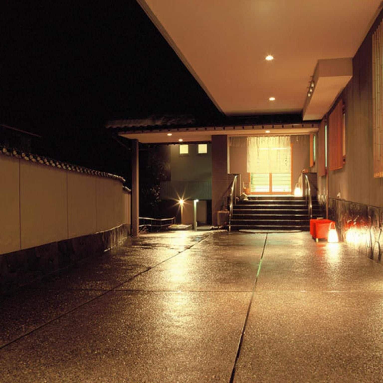 A nighttime view of a building's exterior with a staircase leading to a door, a wall with a railing, and a light source above the door.