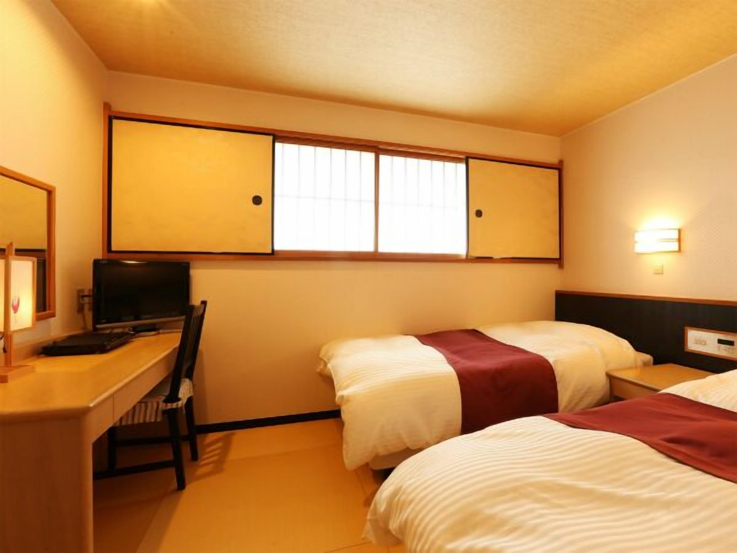 A hotel room with a desk, two beds, a television, and a window with blinds.