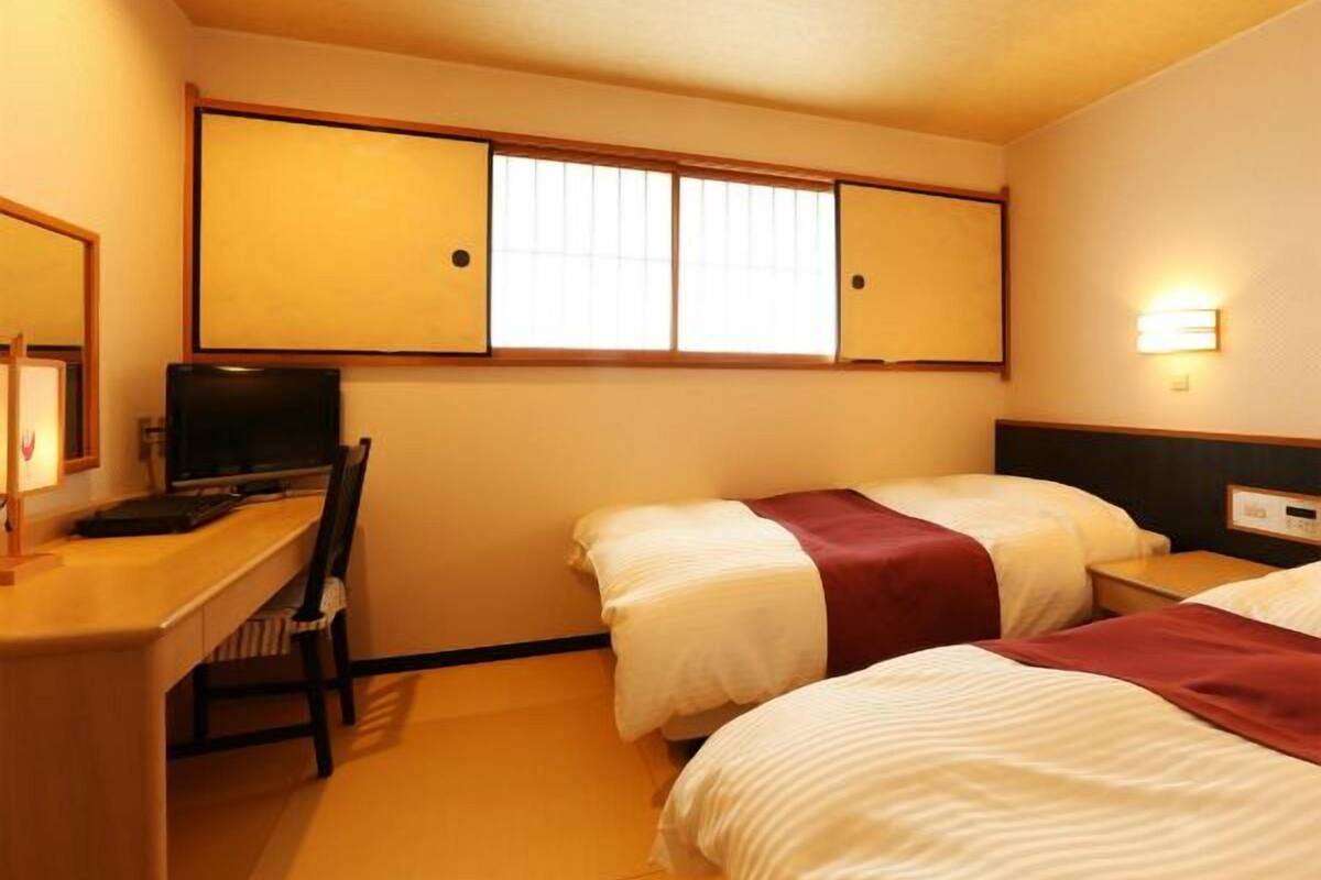 A hotel room with a desk, two beds, a television, and a window with blinds.
