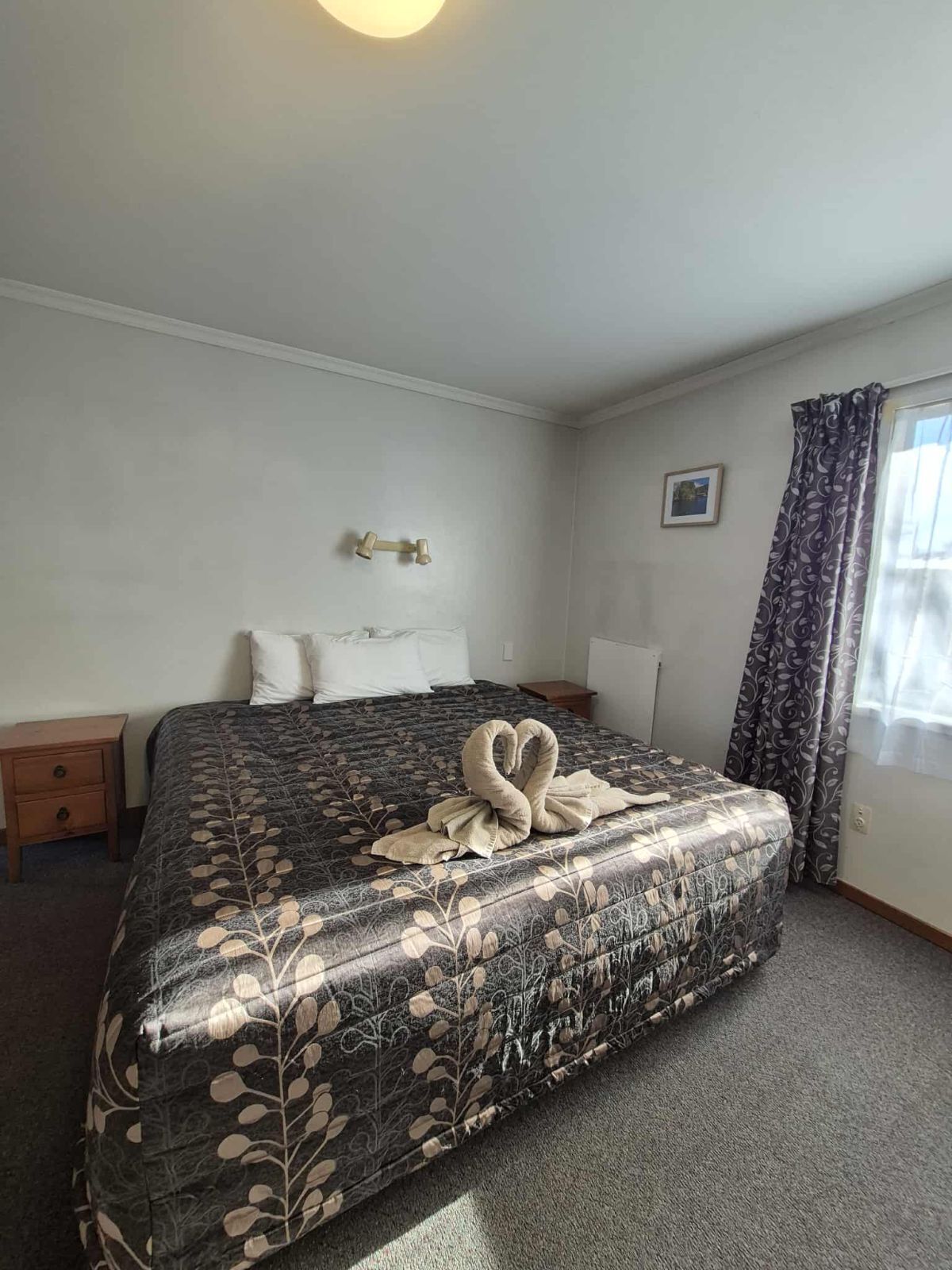 A hotel room with a bed, a nightstand, a lamp, a picture on the wall, and a window with curtains.