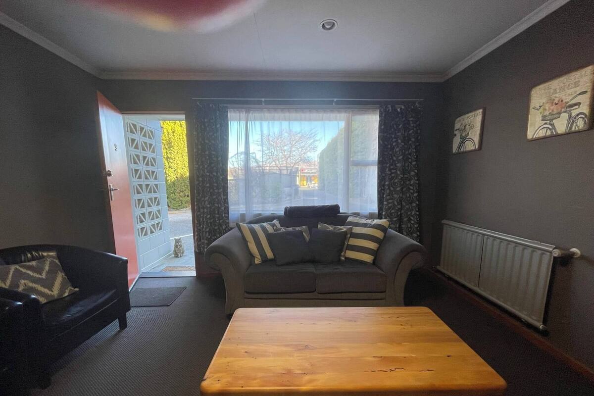 Una sala de estar con un sofá, una mesa de centro de madera y una silla. Se ve una ventana con cortinas, un radiador y una puerta que da al exterior.