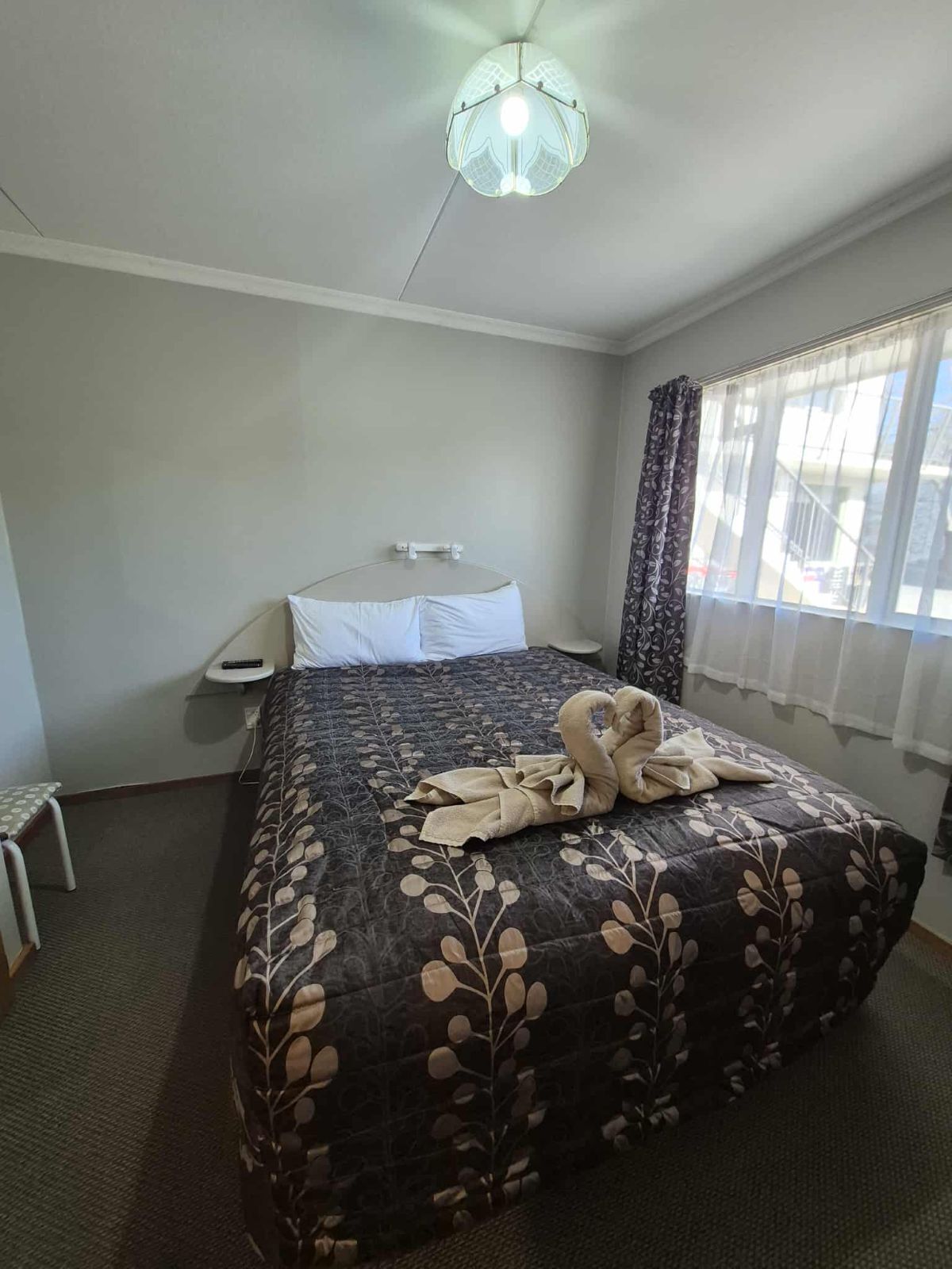 Quarto de hotel com cama, uma pequena mesa, uma cadeira, janela com cortinas e luminária no teto.