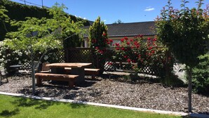 Un patio con mesa y bancos de madera, rodeado de setos y plantas con flores.