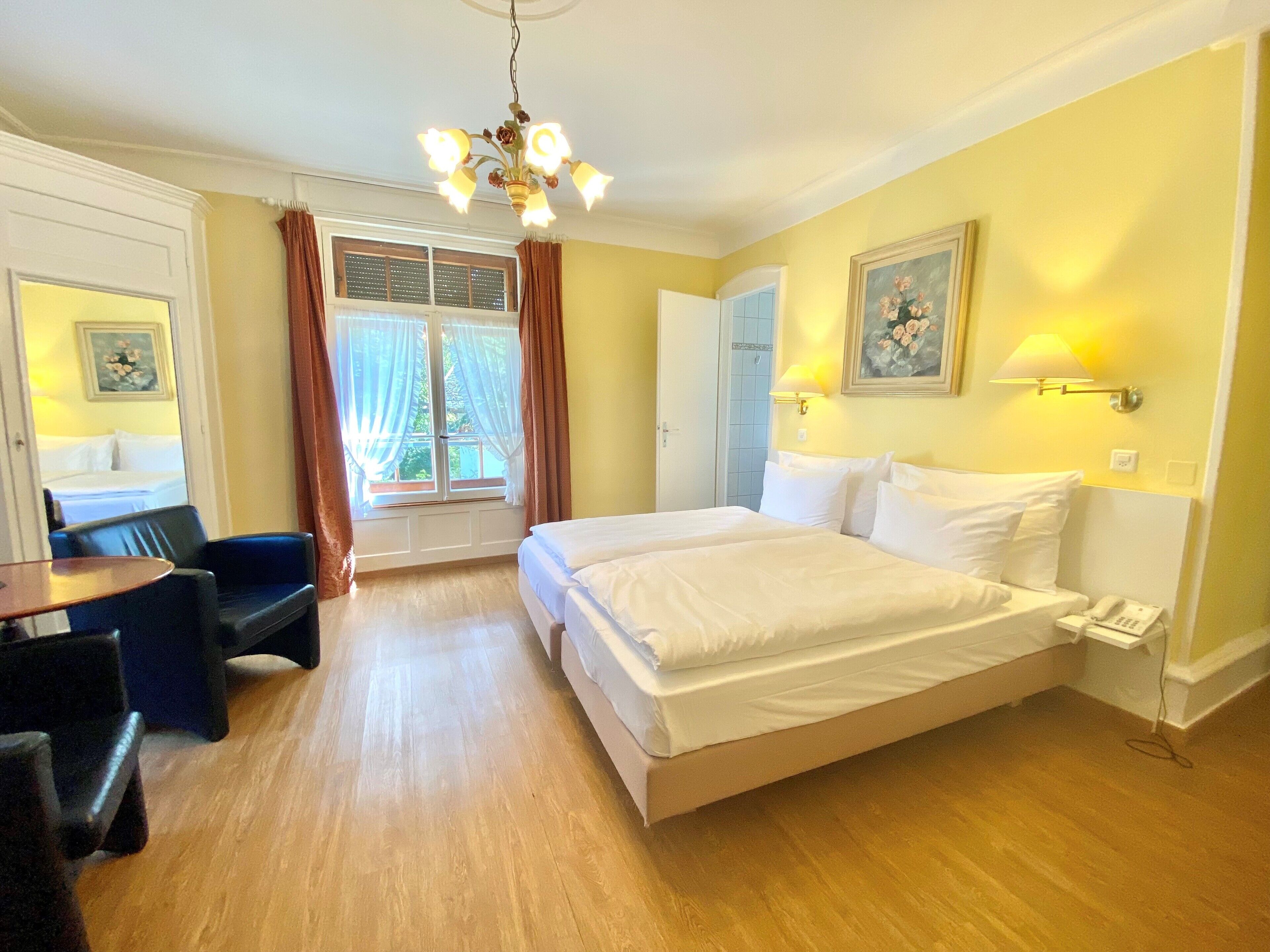 A hotel room with a bed, a desk, a chair, a window with curtains, a chandelier, and a painting on the wall.