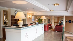 A reception area with a white counter, two lamps, a clock, and a plant. - Hôtel La Prairie, Swiss Bike Hotels (Yverdon-les-Bains)