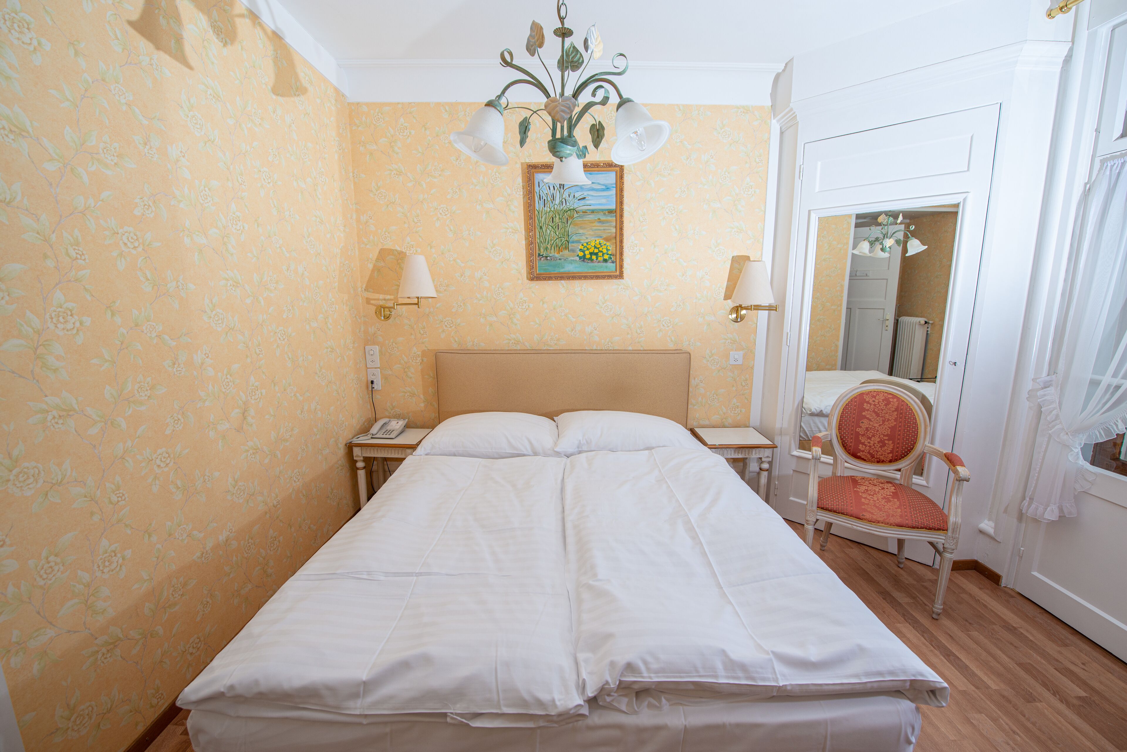 A neatly made bed in a room with floral wallpaper, a painting on the wall, and a chair with a red cushion.