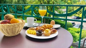 A breakfast table with a wicker basket of fruits, a plate of pastries, a glass of orange juice, and a cup on a balcony overlooking a parking area and greenery. - Hôtel La Prairie, Swiss Bike Hotels (Yverdon-les-Bains)