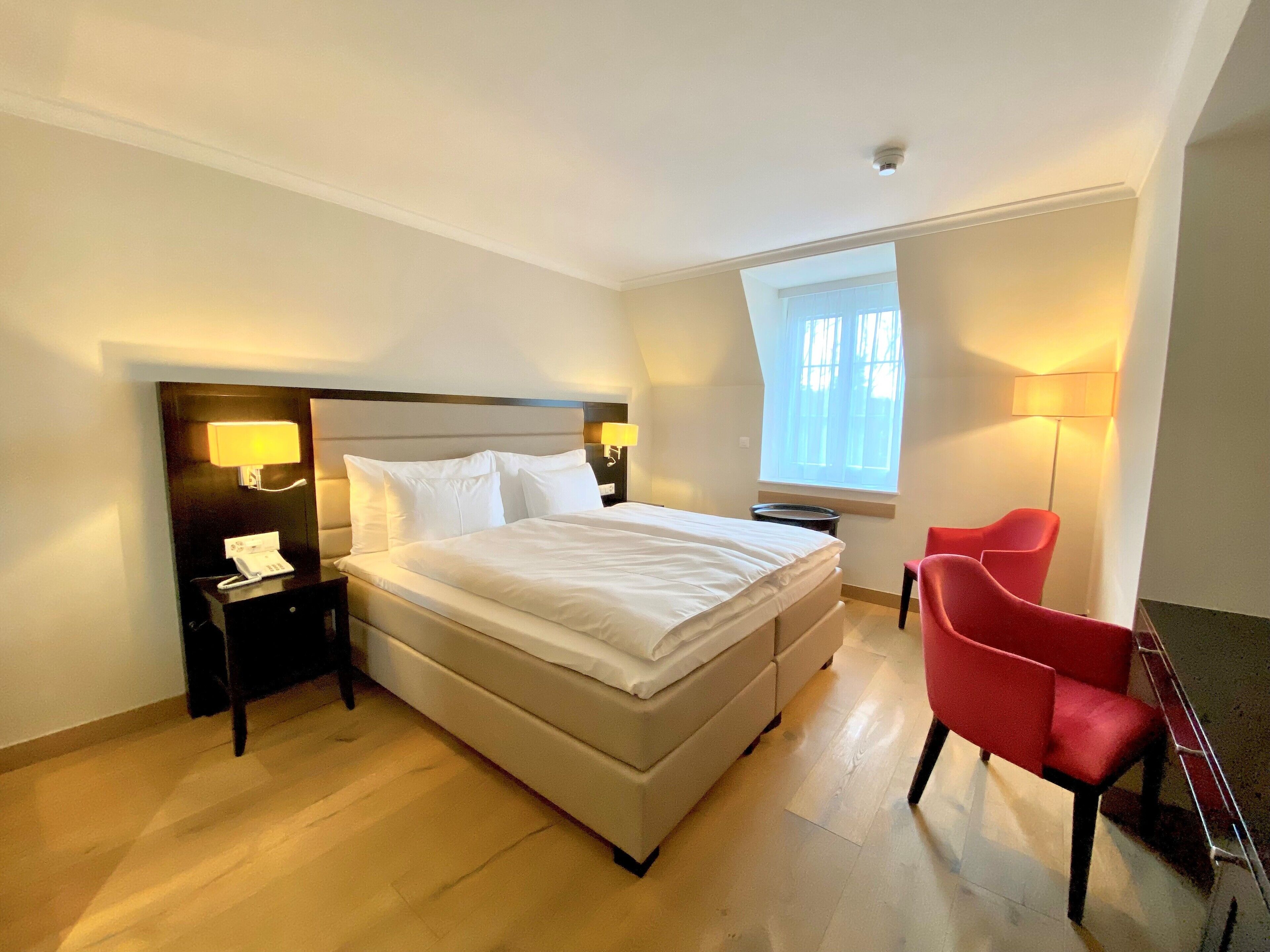 A hotel room with a large bed, bedside lamps, a red chair, and a window with natural light.