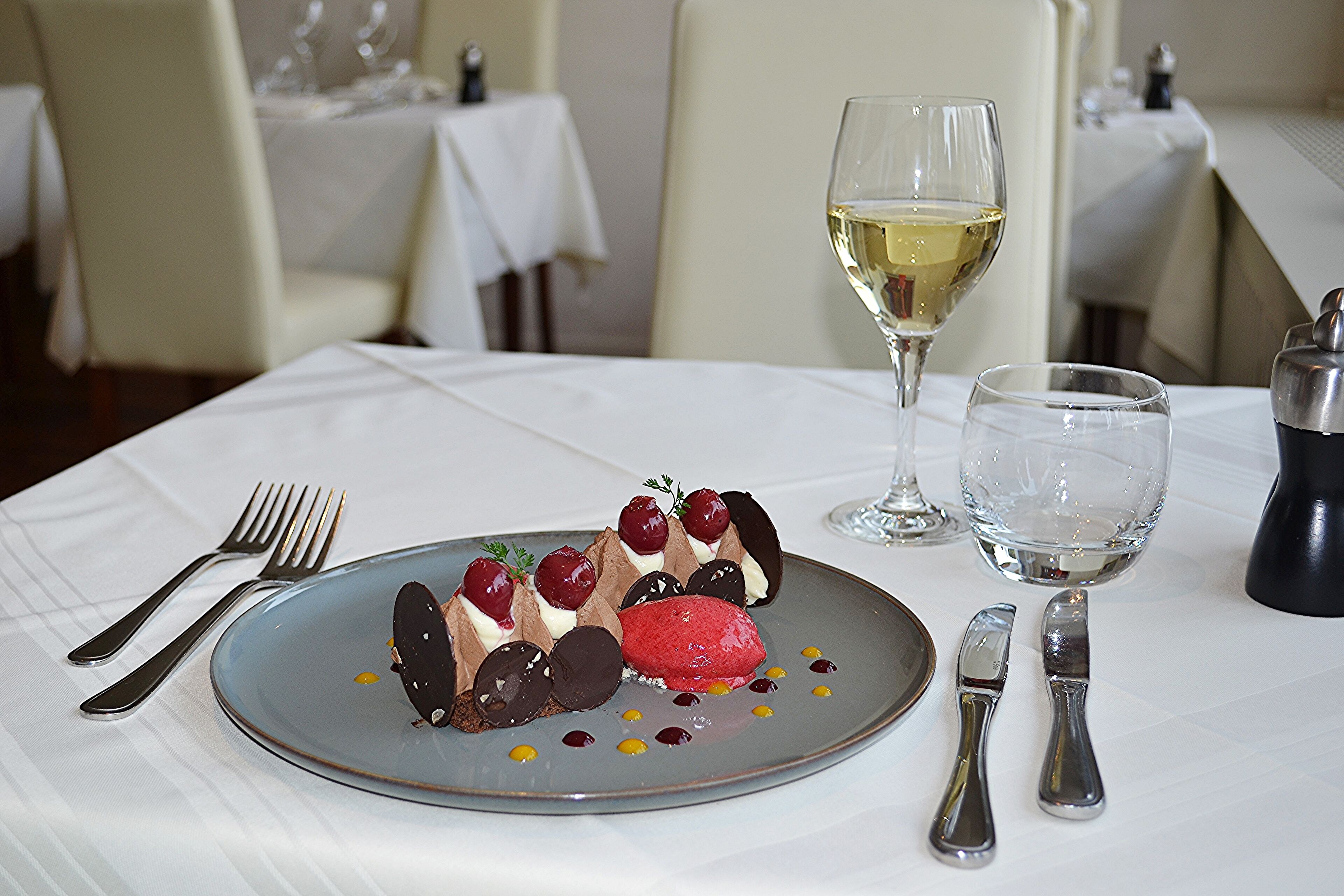 Une assiette à dessert contenant un dessert au chocolat et aux cerises, accompagnée d’un verre de vin blanc et d’un verre d’eau, le tout posé sur une table dans un restaurant.