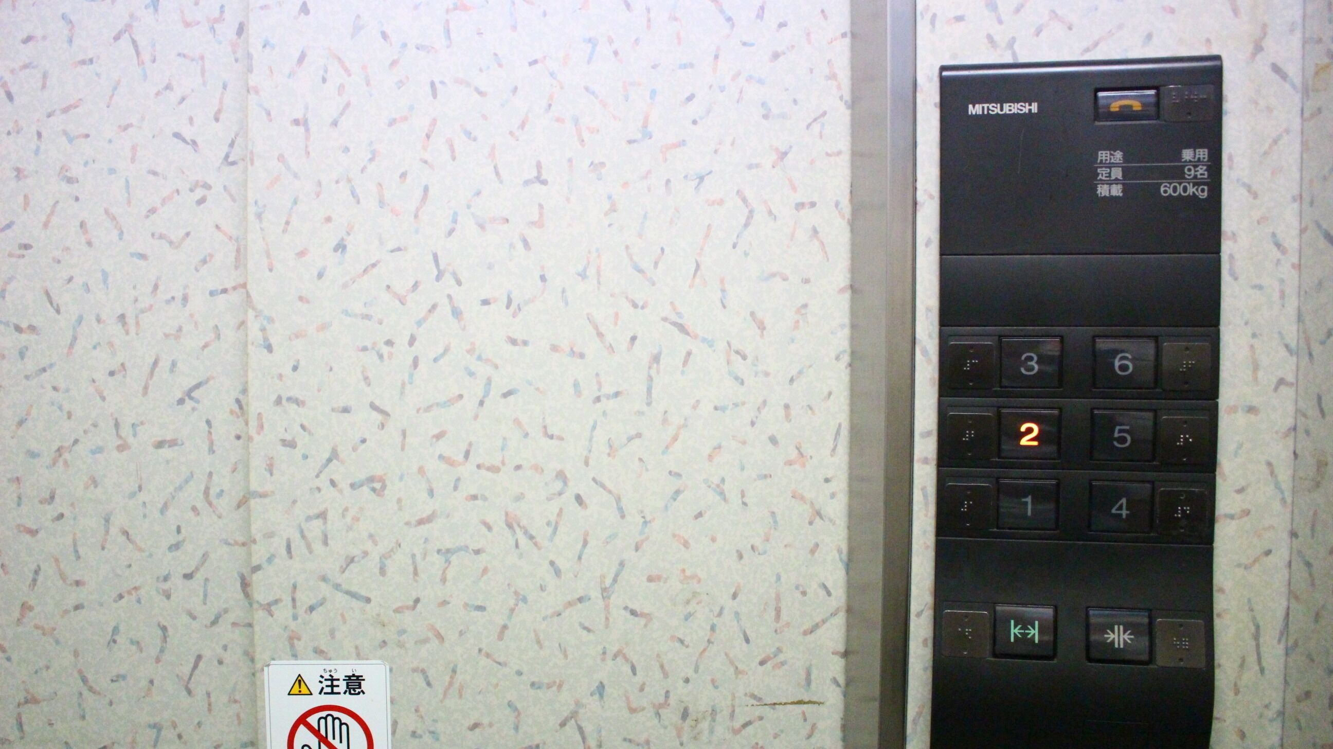 An elevator interior with a Mitsubishi brand control panel, a warning sign, and a floor indicator.