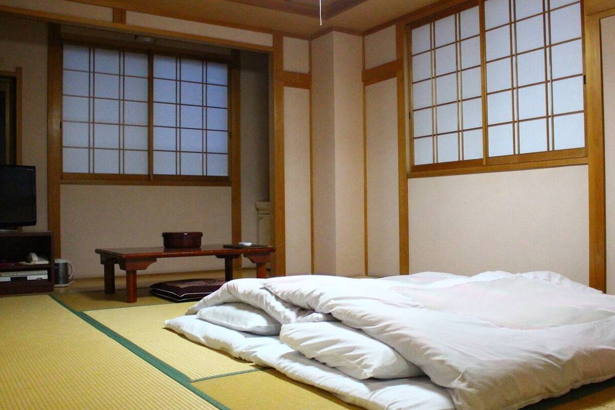 Une pièce de style japonais traditionnel, avec un sol recouvert de tatamis, un lit avec une literie blanche, un téléviseur posé sur un meuble et des portes coulissantes.