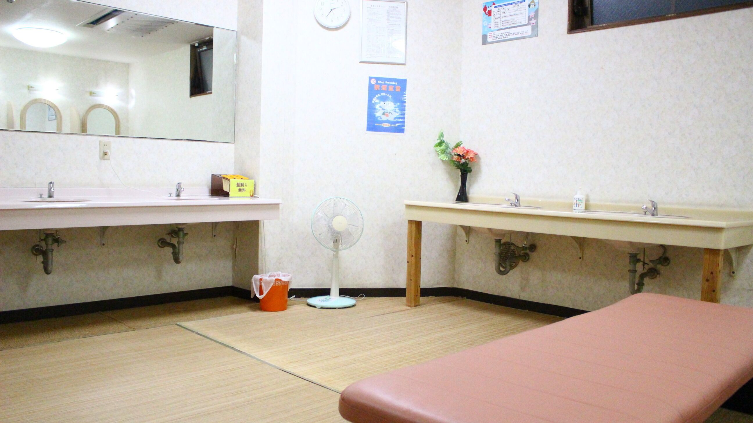 A room with two sinks, a mirror, a clock, a fan, a trash bin, and a small table with a vase.