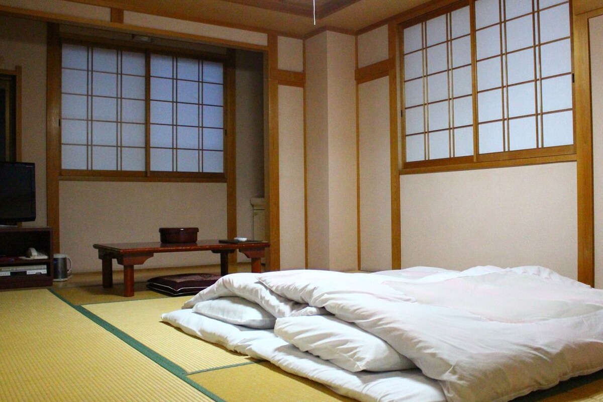 A traditional Japanese room with tatami flooring, a bed with white bedding, a television on a stand, and sliding doors.